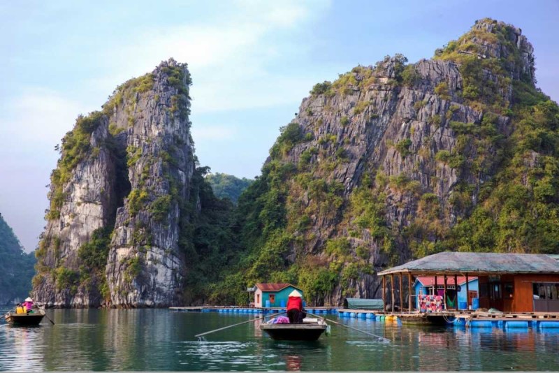 Bamboo boat navigating calm waters near floating homes Cua Van Bamboo boat rowed by a local woman near floating homes at Cua Van Floating Village, Halong Bay with dramatic karst cliffs behind