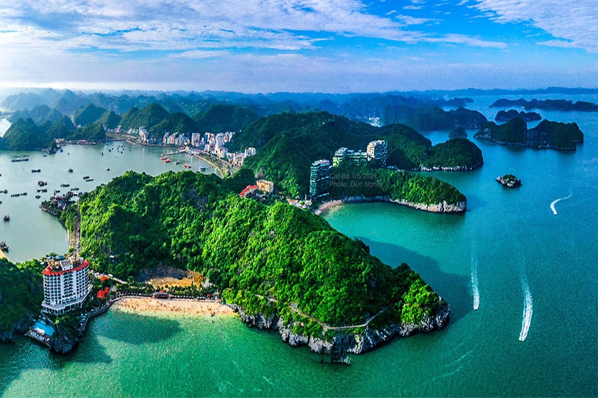 A breathtaking aerial view of Cat Ba Island in Halong Bay, showcasing lush green hills, turquoise waters, and coastal towns. Boats are seen moving across the bay, offering a glimpse into the serene beauty of this hidden gem.