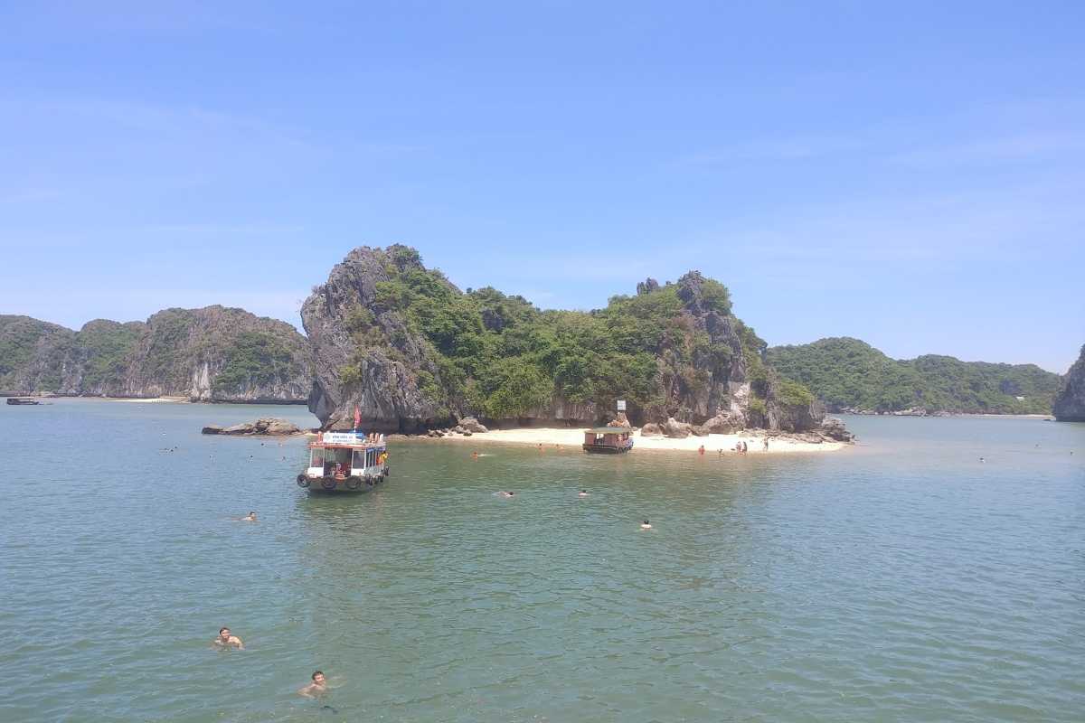 Ba Trai Dao Beach Halong Bay boats anchored near sandy shore Boats anchored at Ba Trai Dao Beach Halong Bay with swimmers and clear skies surrounded by lush islets.