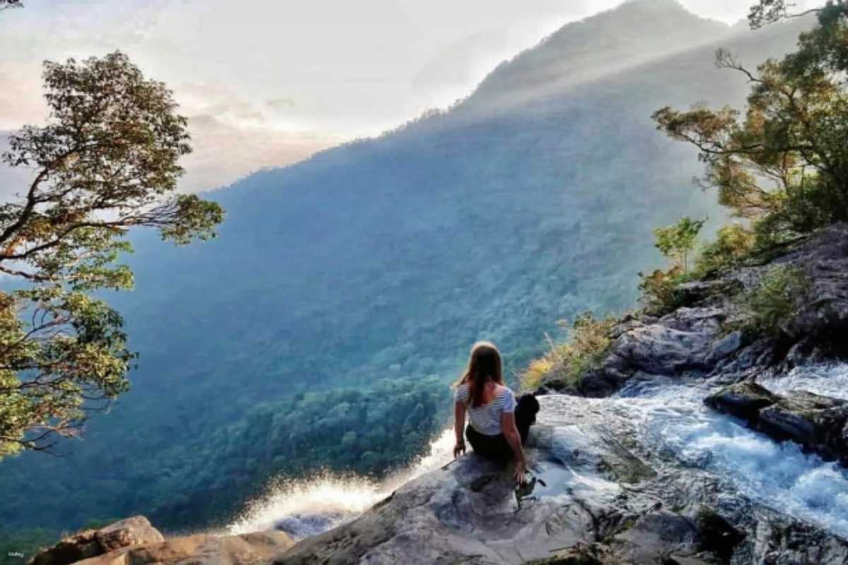 Relaxing at Waterfall Edge with Mountain Views Woman sitting at the edge of a cascading waterfall overlooking dense green mountains in Bach Ma National Park