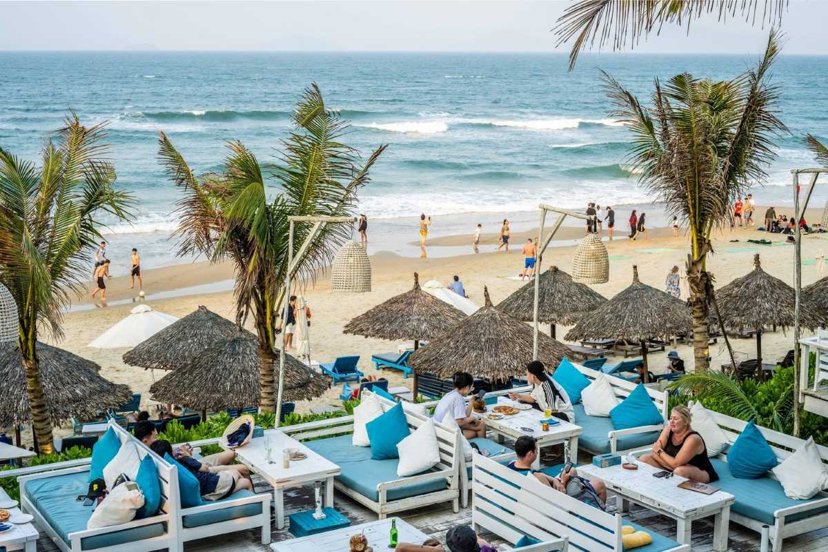 Beachfront Dining at An Bang Beach Relaxed guests dining at white tables with blue cushions under palm trees by the sandy beach