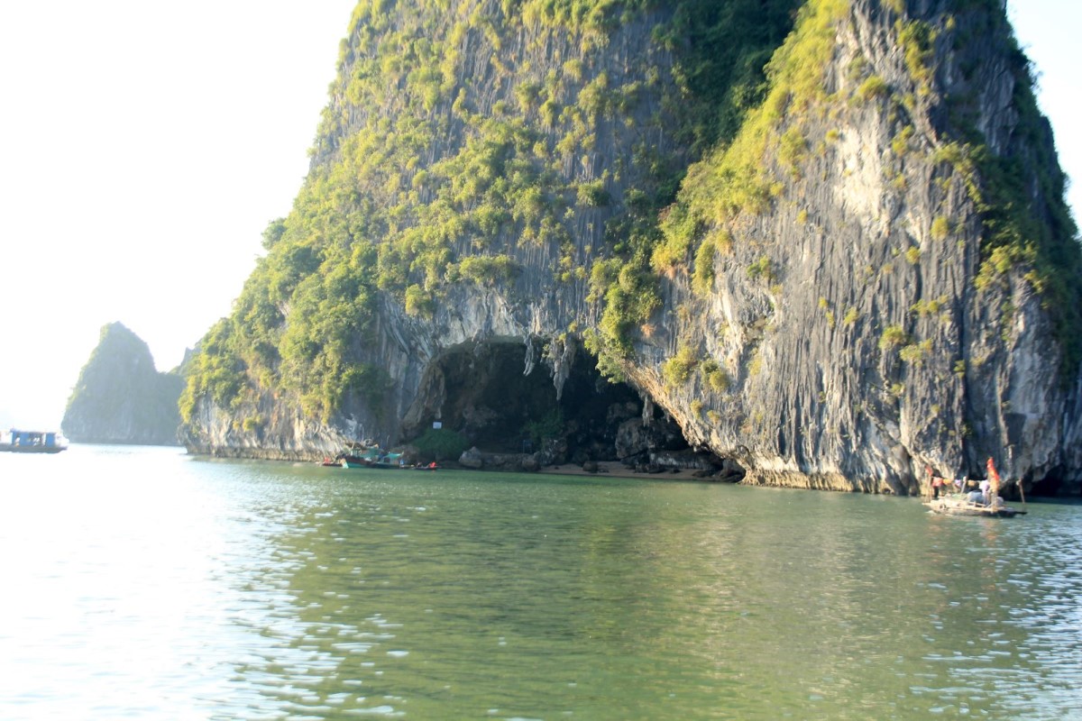 Entrance of Bo Nau Cave surrounded by towering limestone cliffs in Halong Bay, with calm emerald waters in the foreground.