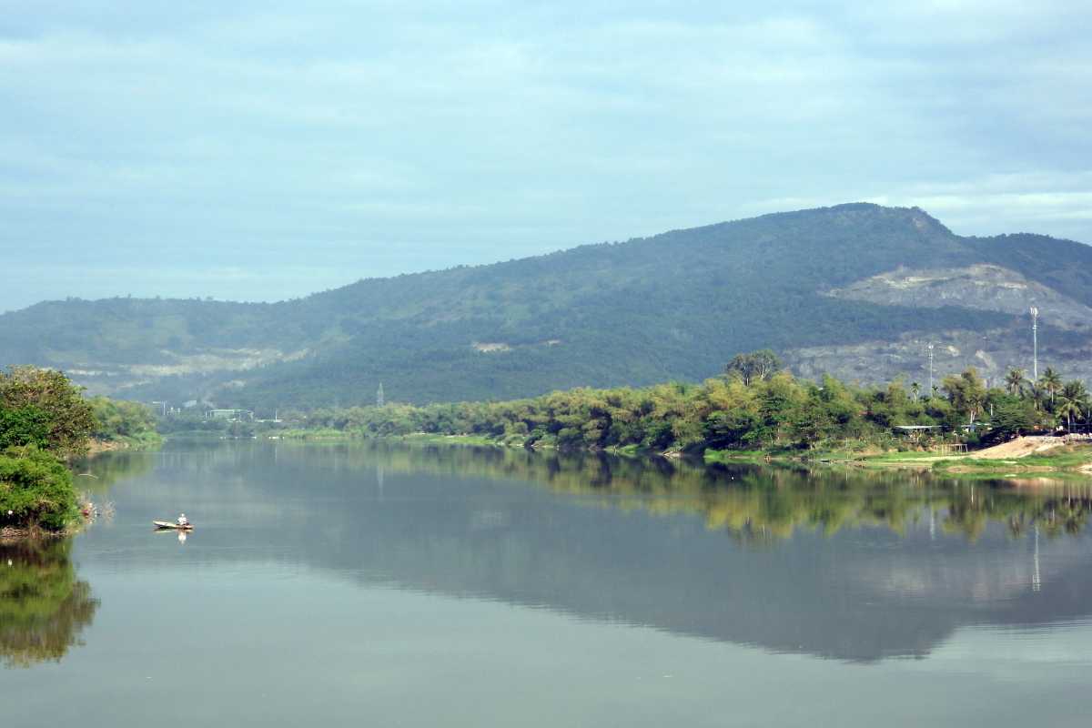Forest-Framed Tranquil Section of Cai River A calm, reflective stretch of the Cai River lined with dense trees and distant mountains under a cloudy sky.