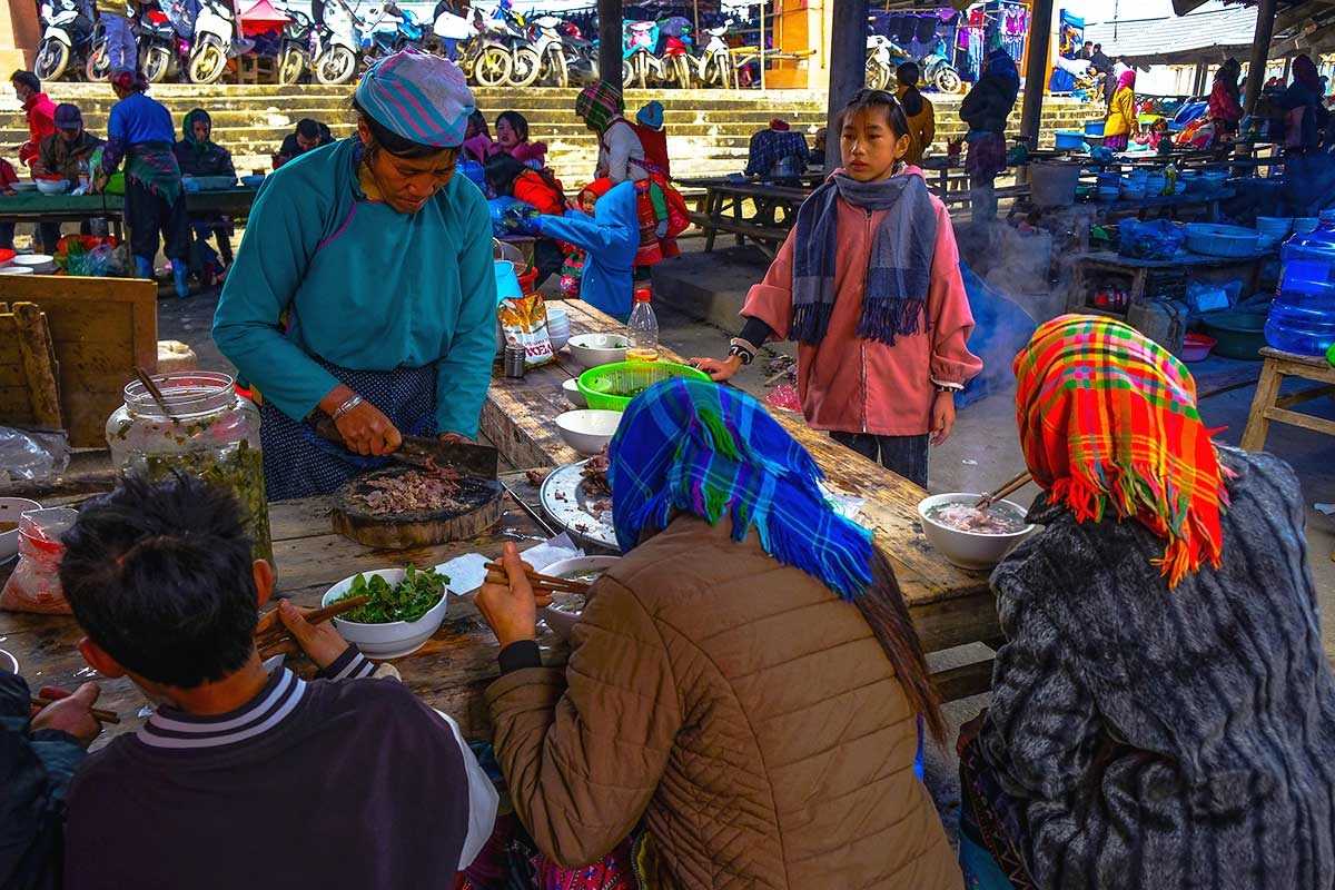 Can Cau Market Lao Cai food stall with ethnic diners A local woman serves grilled meat while ethnic diners enjoy hot food at Can Cau Market’s rustic food stall