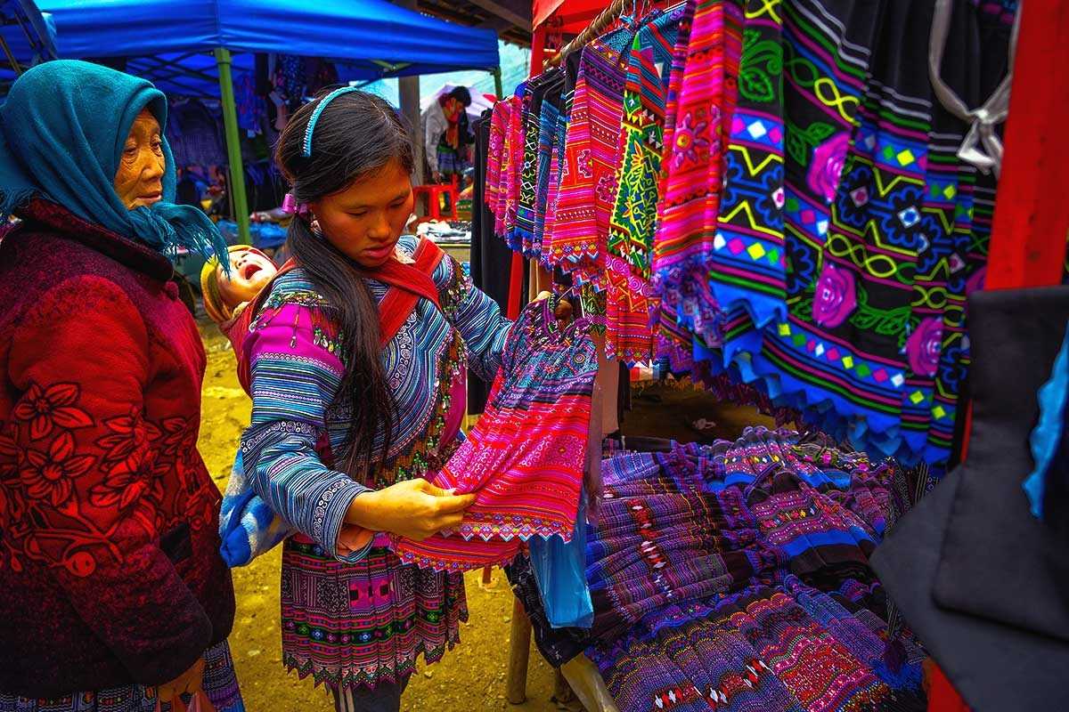Can Cau Market Lao Cai woman browsing traditional textiles A woman with baby browses vibrant traditional Hmong textiles at a Can Cau Market stall in Lao Cai