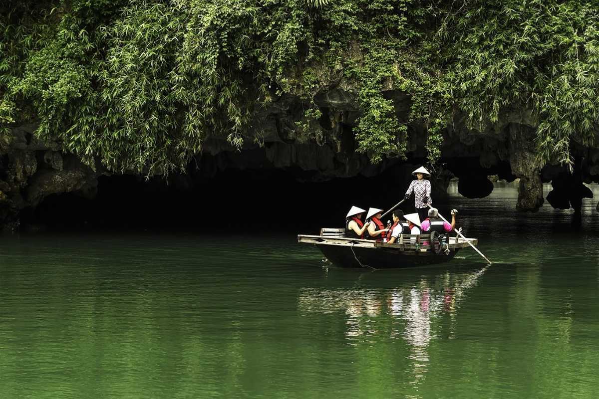 Dark and Bright Cave Halong Bay rowboat entrance greenery Dark and Bright Cave Halong Bay local rowboat with tourists approaches cave entrance surrounded by lush green foliage.