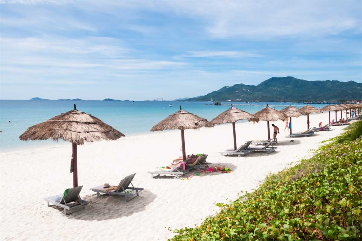 Beachfront view of Doc Let Beach with palapa umbrellas, wooden lounge chairs, and clear turquoise water under a sunny sky.