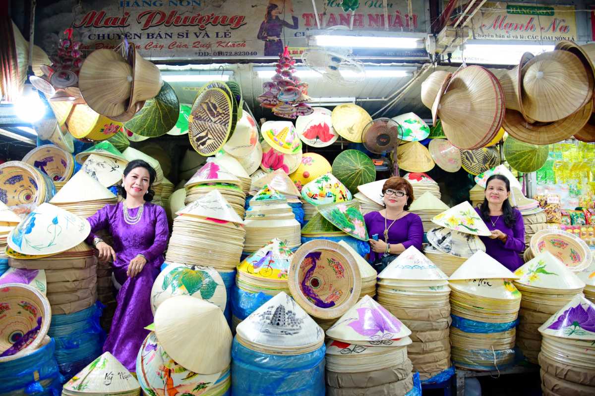 Women in Ao Dai at Dong Ba Market Conical Hat Stalls Three women in purple Ao Dai surrounded by stacks of Non Bai Tho conical hats at Dong Ba Market Hue