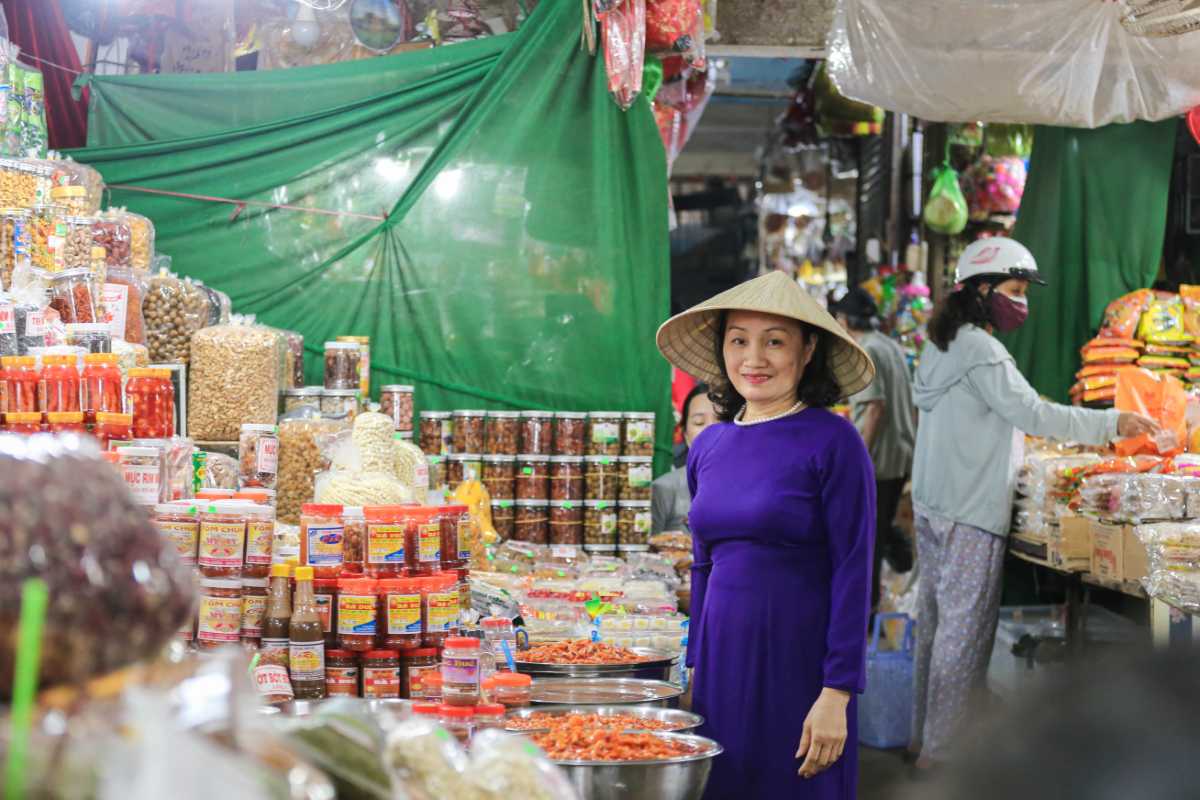 Dong Ba Market Vendor in Ao Dai with Local Specialties A Dong Ba Market vendor in purple Ao Dai and conical hat stands by colorful jars of Hue specialties