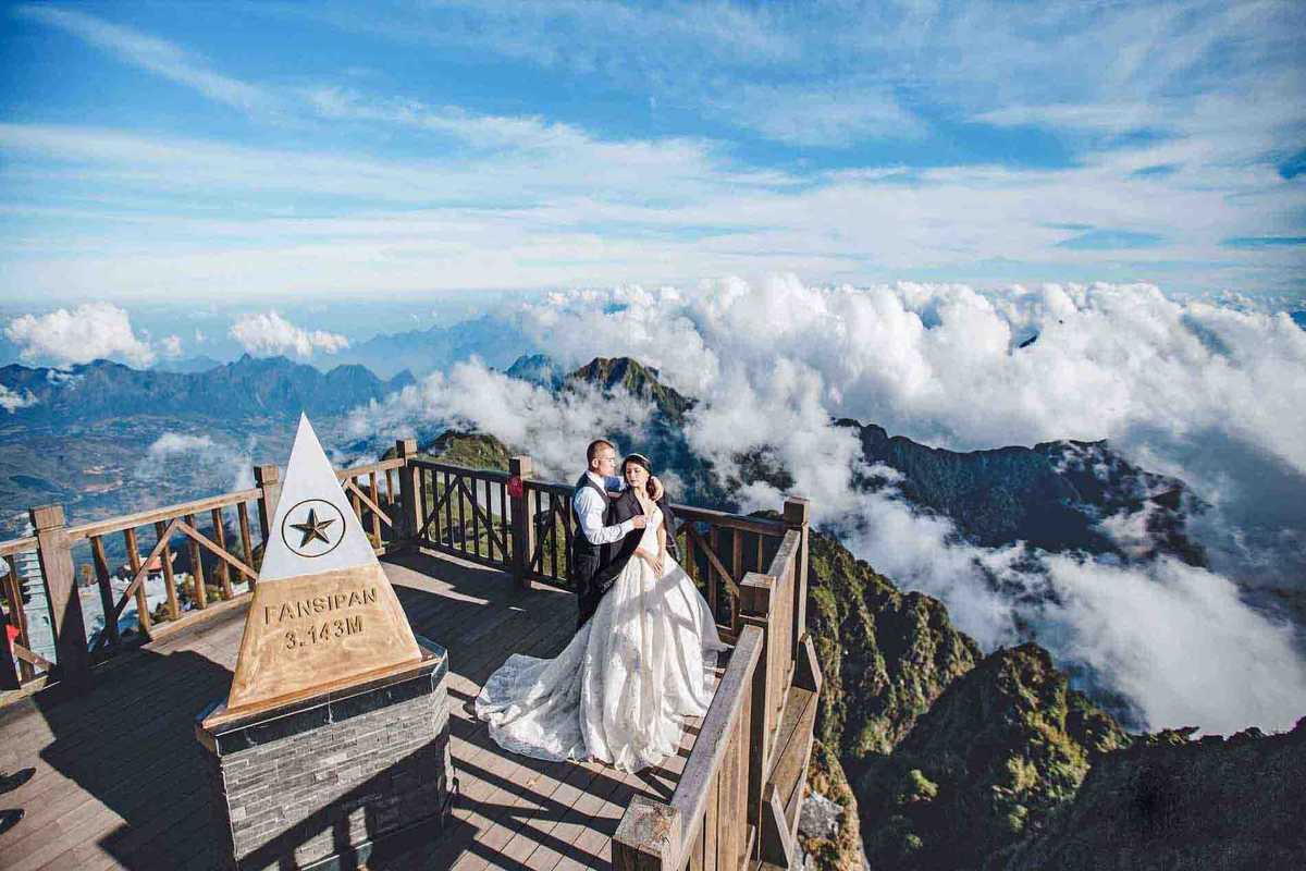 Fansipan Mountain Sapa summit wedding photography spot Fansipan Mountain Sapa summit marker with newlywed couple posing above clouds and rugged mountain peaks