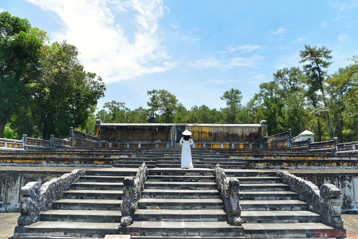 Dragon Staircase with Visitor in Ao Dai Woman in white ao dai standing atop the dragon-carved stone staircase at Gia Long Tomb in Hue, surrounded by trees.