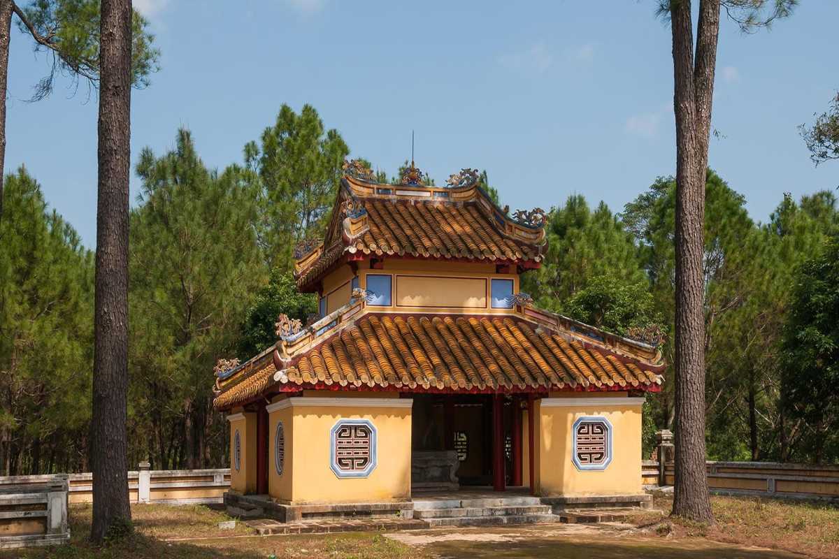 Pine-Shaded Pavilion at Gia Long Tomb Small yellow pavilion with curved tiled roof, surrounded by tall pines inside Gia Long Tomb complex in Hue, Vietnam.