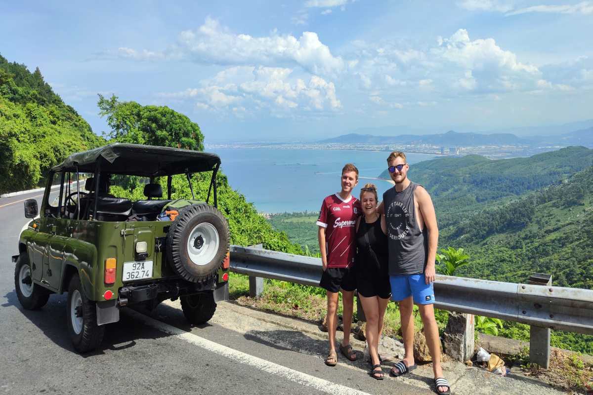 Hai Van Pass jeep tour travelers Da Nang bay backdrop Hai Van Pass jeep tour travelers pose above Da Nang Bay, with a scenic mountain backdrop and coastal panorama.