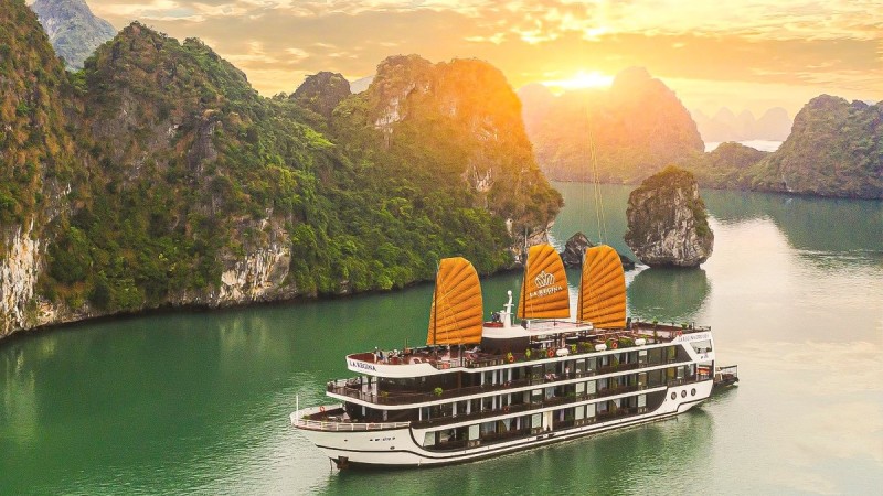 A luxurious overnight cruise boat sails through Halong Bay at sunset, with the towering cliffs of Titov Island in the distance.