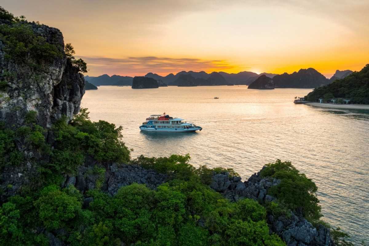 A sunset cruise boat floats on tranquil Halong Bay waters surrounded by limestone cliffs and a glowing sky.