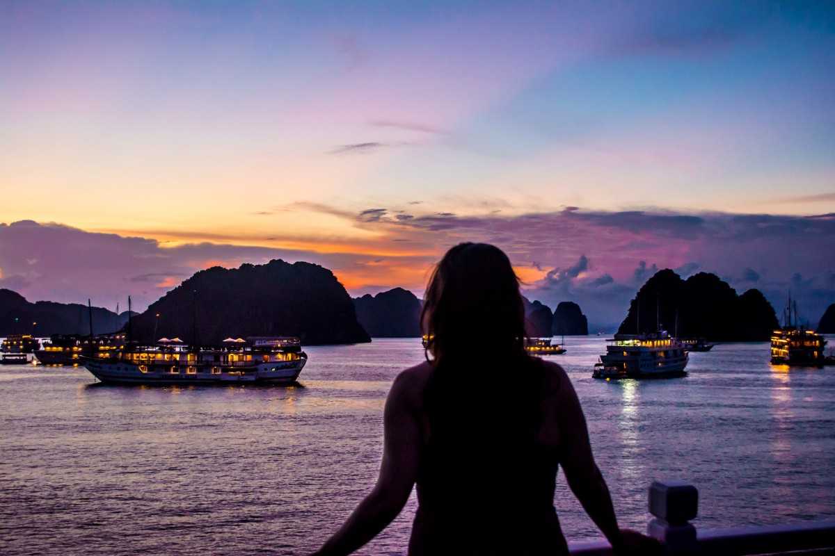Panoramic view of Halong Bay sunset with tourist A traveler watches the vivid Halong Bay sunset from a cruise deck surrounded by anchored boats and islets.