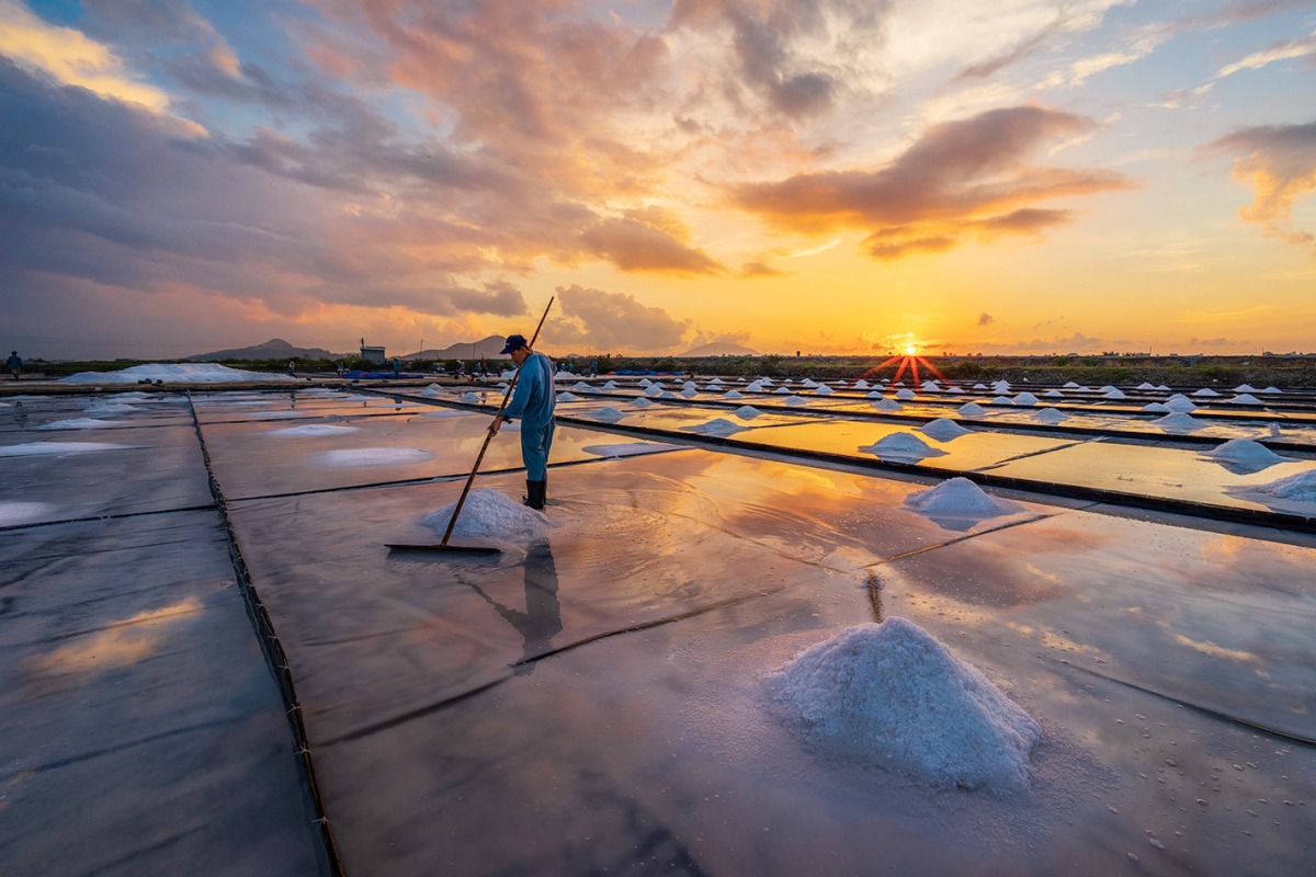 Salt fields in Hon Khoi Commune reflecting a vibrant sunset sky with workers harvesting salt
