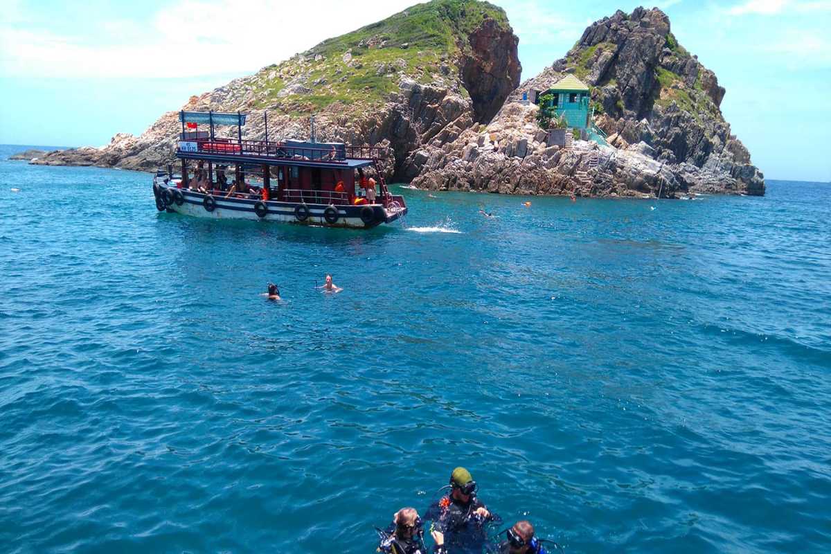 Snorkeling boat and swimmers at Hon Mun Island Tour boat and tourists swimming near a rocky cliff area of Hon Mun Island under bright skies.
