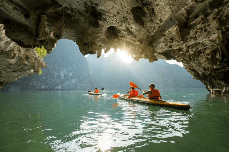 Kayakers paddling through a scenic cave in Halong Bay, with sunlight streaming through the limestone formations near Titov Island.