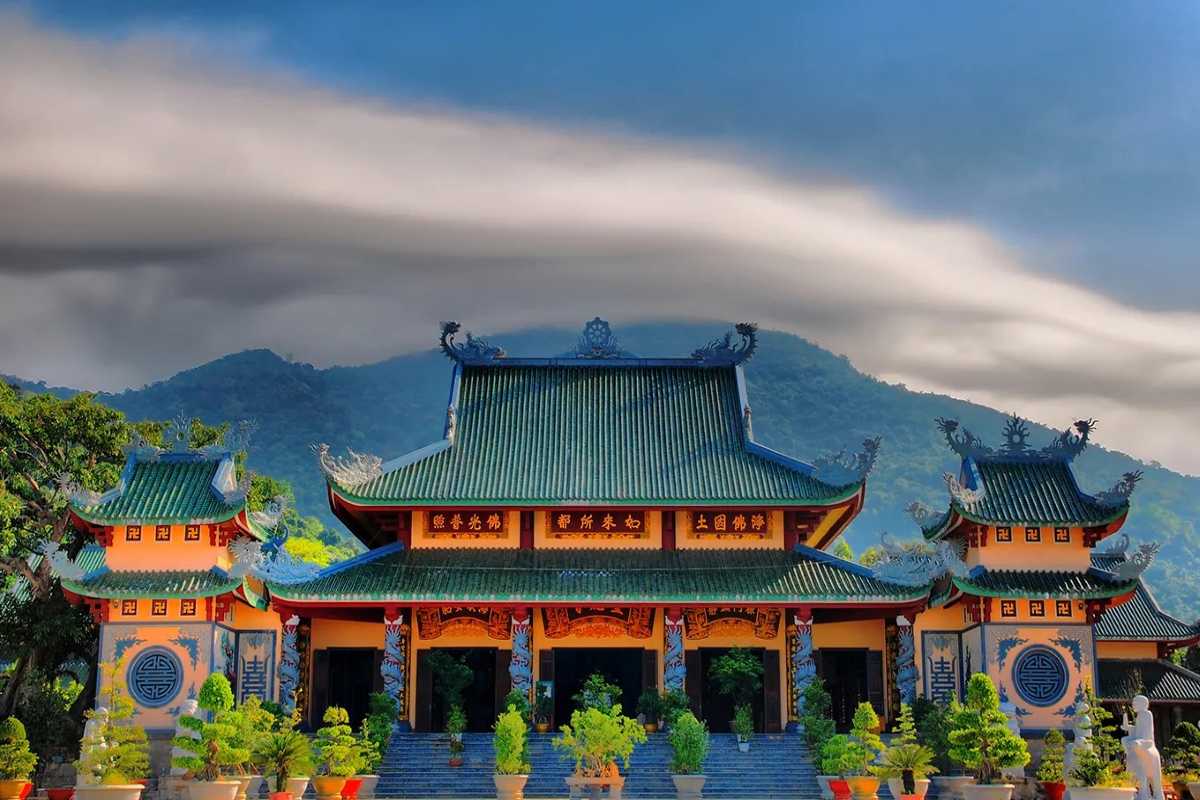 Linh Ung Pagoda Da Nang main hall with green tiled roof Linh Ung Pagoda Da Nang main hall with green-tiled roof, ornate woodwork, and mountains rising in the background under dramatic sky.