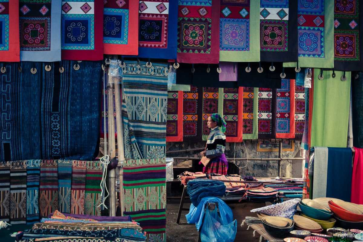 Lung Khau Nhin Market brocade shop with colorful textiles A local vendor at Lung Khau Nhin Market arranges vivid brocade panels featuring H'mong motifs and traditional designs