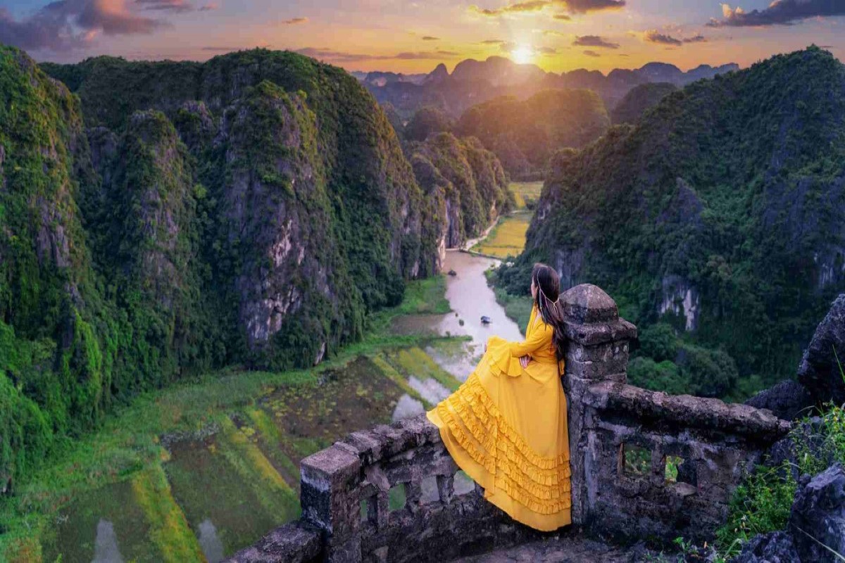 A breathtaking view of the sunset over Mua Caves in Ninh Binh, with a woman in a yellow dress overlooking the lush green valley below.