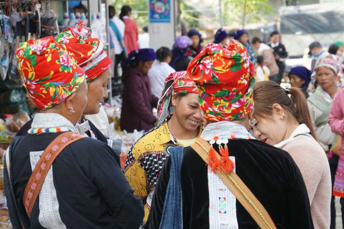 Muong Hum Market Sapa Red Dao women lively marketplace interaction Red Dao women in traditional attire chatting and interacting at Muong Hum Market Sapa’s lively open-air stalls.