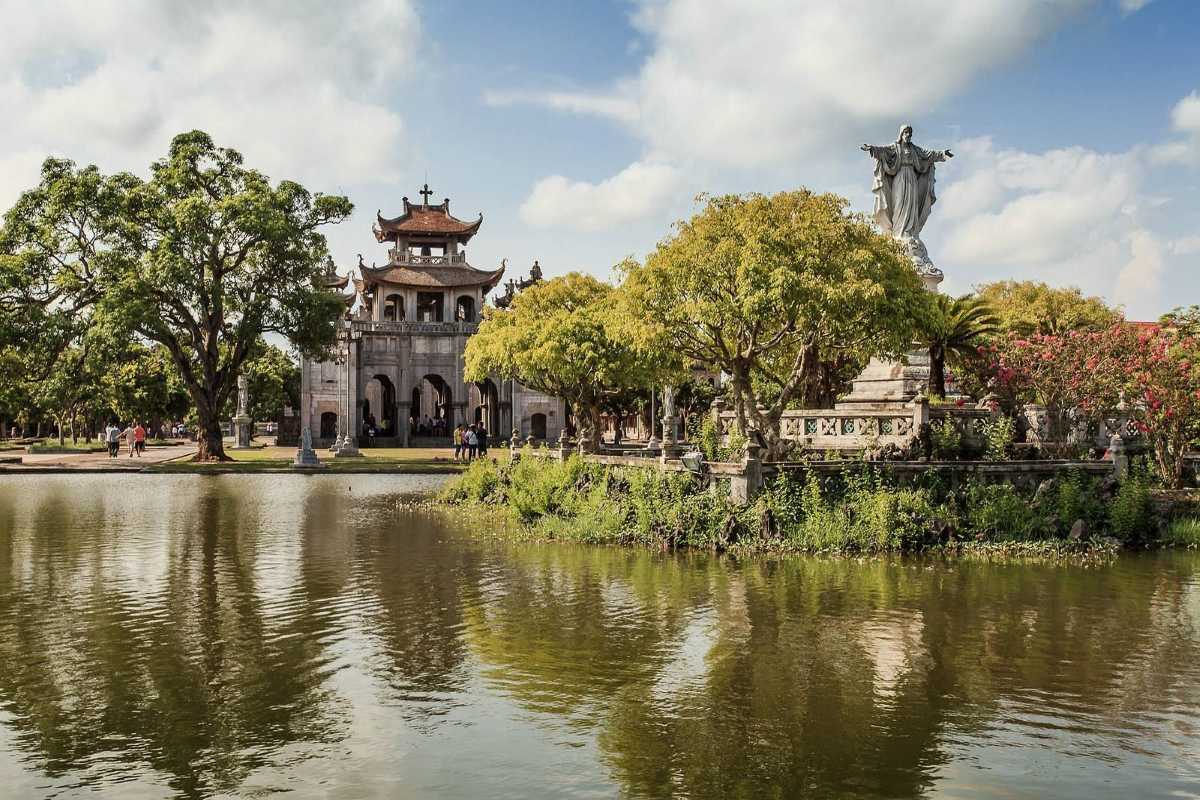 Phat Diem Cathedral Ninh Binh lakeside view with statue Phat Diem Cathedral Ninh Binh viewed from across a tranquil lake, framed by trees and a prominent Jesus statue in the garden.
