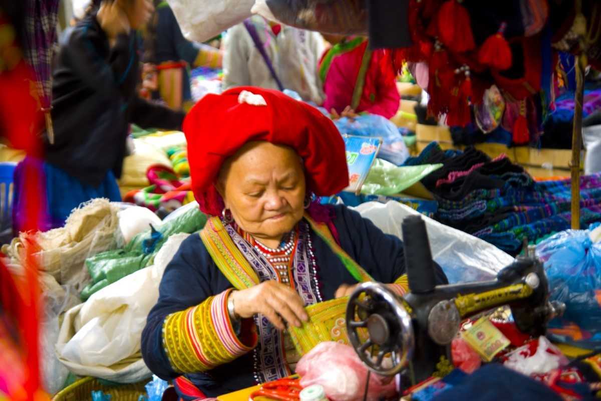 Red Dzao woman sewing at Coc Ly Market An elderly Red Dzao woman concentrates on sewing at her fabric stall inside Coc Ly Market, surrounded by colorful threads.