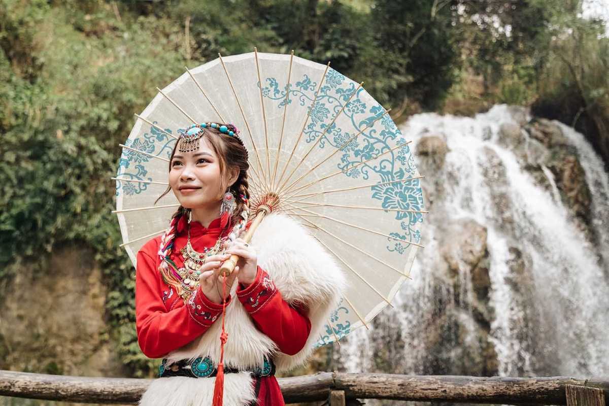 Sapa Love Market Vietnam Hmong girl cultural attire Sapa Love Market Vietnam displays a young Hmong girl in traditional dress with a parasol, standing by a waterfall.