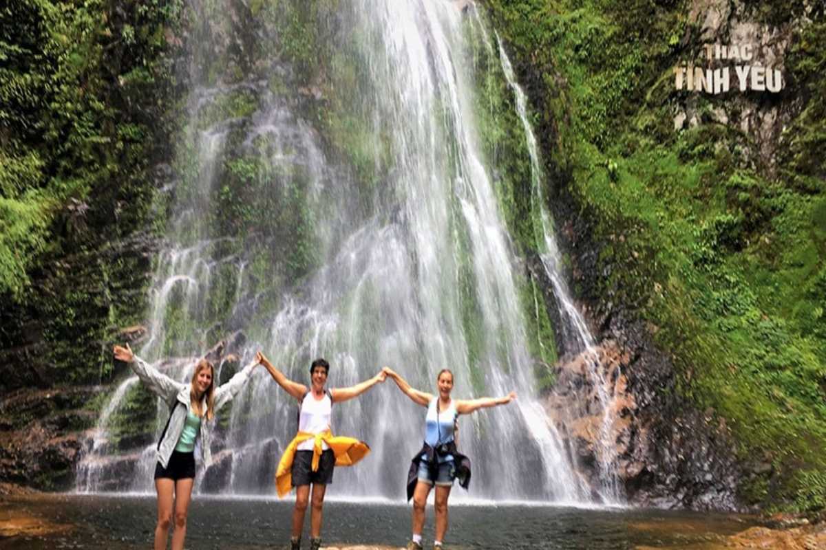 Sapa Love Waterfall Sapa joyful hikers at the falls Three happy hikers raise their arms in front of the majestic Sapa Love Waterfall, framed by lush greenery and falling water.