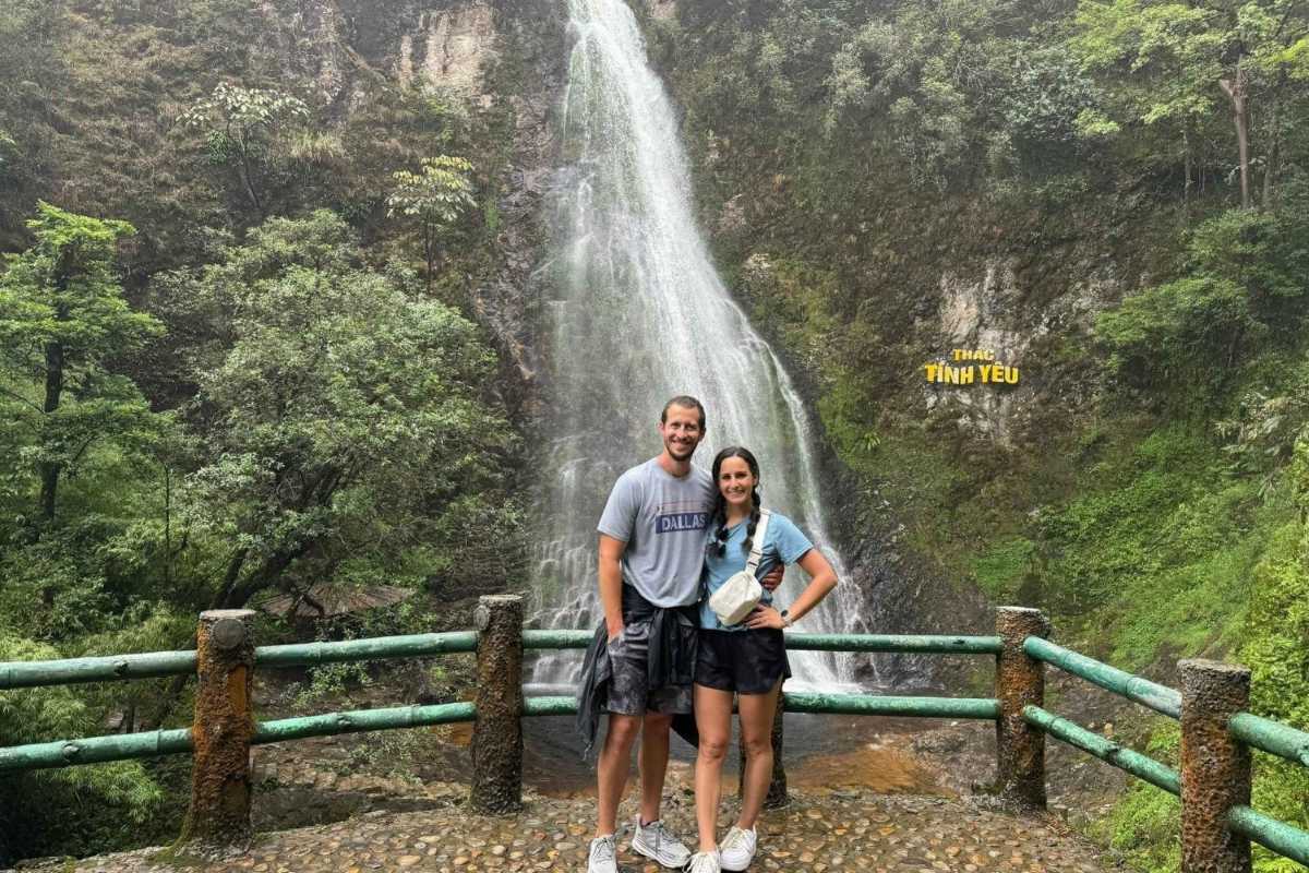 Sapa Love Waterfall Sapa scenic couple viewpoint A couple stands in front of Sapa Love Waterfall at a popular viewpoint, enjoying the vibrant greenery and falling water.