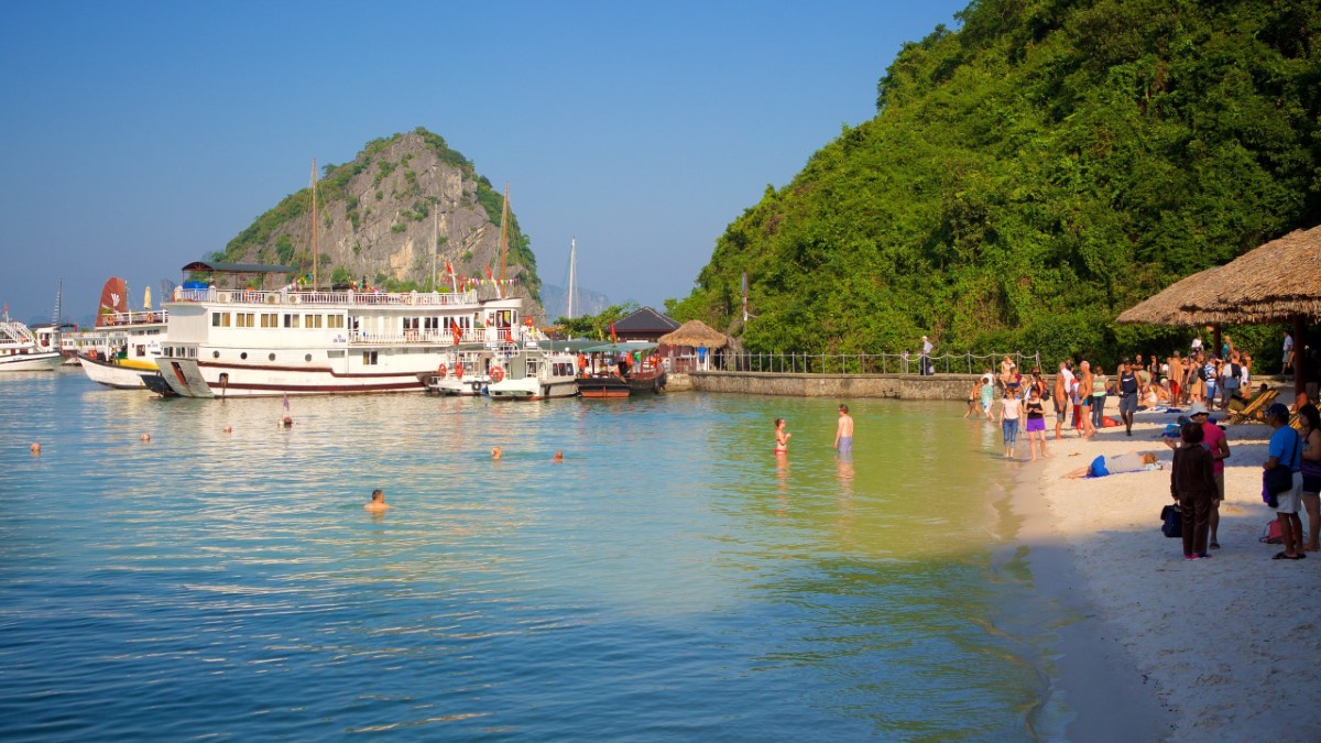 Soi Sim Island beach with tourist boats docked and visitors swimming in Ha Long Bay.