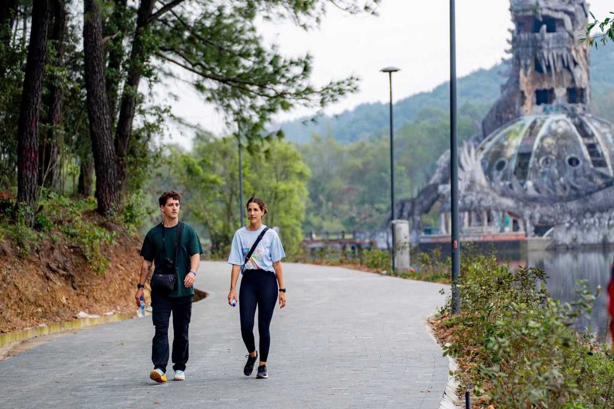 Visitors Strolling Path at Thuy Tien Lake Abandoned Water Park Two travelers walk on a lakeside path, with the dragon dome of Thuy Tien Lake Abandoned Water Park in the background