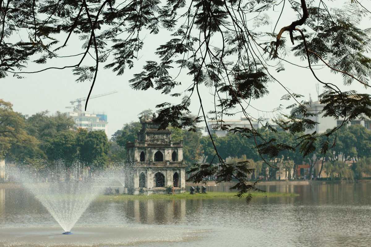 Turtle Tower by Daylight with City Background Daytime view of Turtle Tower with Hanoi city buildings in the distance and green foliage
