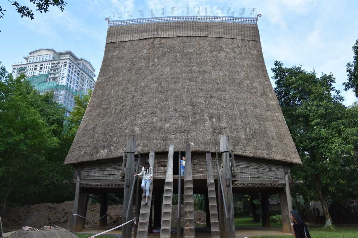 Vietnam Museum of Ethnology Hanoi stilt house visitors experience Visitors exploring traditional stilt house exhibit at Vietnam Museum of Ethnology Hanoi amid lush green surroundings.