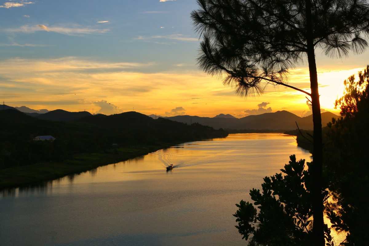 Vong Canh Hill Hue sunset over Perfume River Vong Canh Hill Hue is bathed in golden sunlight at sunset, with the Perfume River shimmering and pine trees silhouetted above.