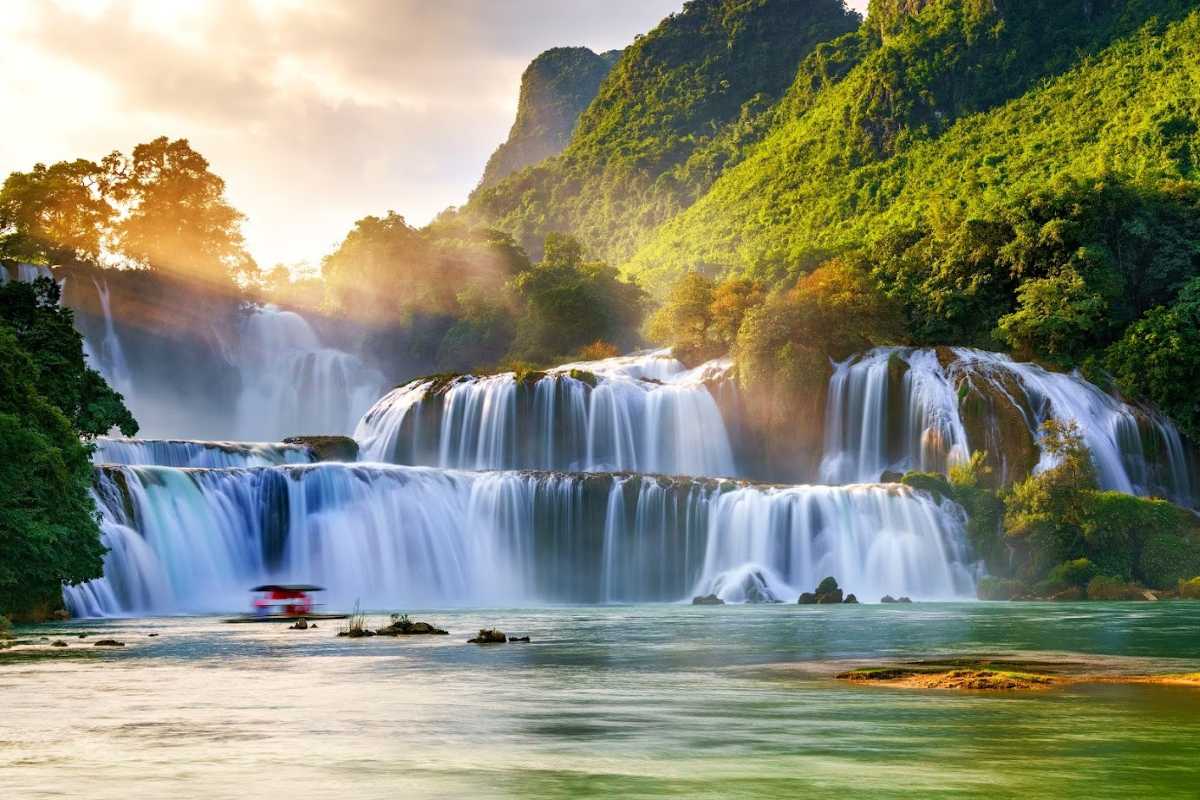 Cao Bang Travel Guide Ban Gioc Waterfall Golden Hour Ban Gioc Waterfall illuminated by golden sunlight with lush greenery and a boat gliding by in Cao Bang