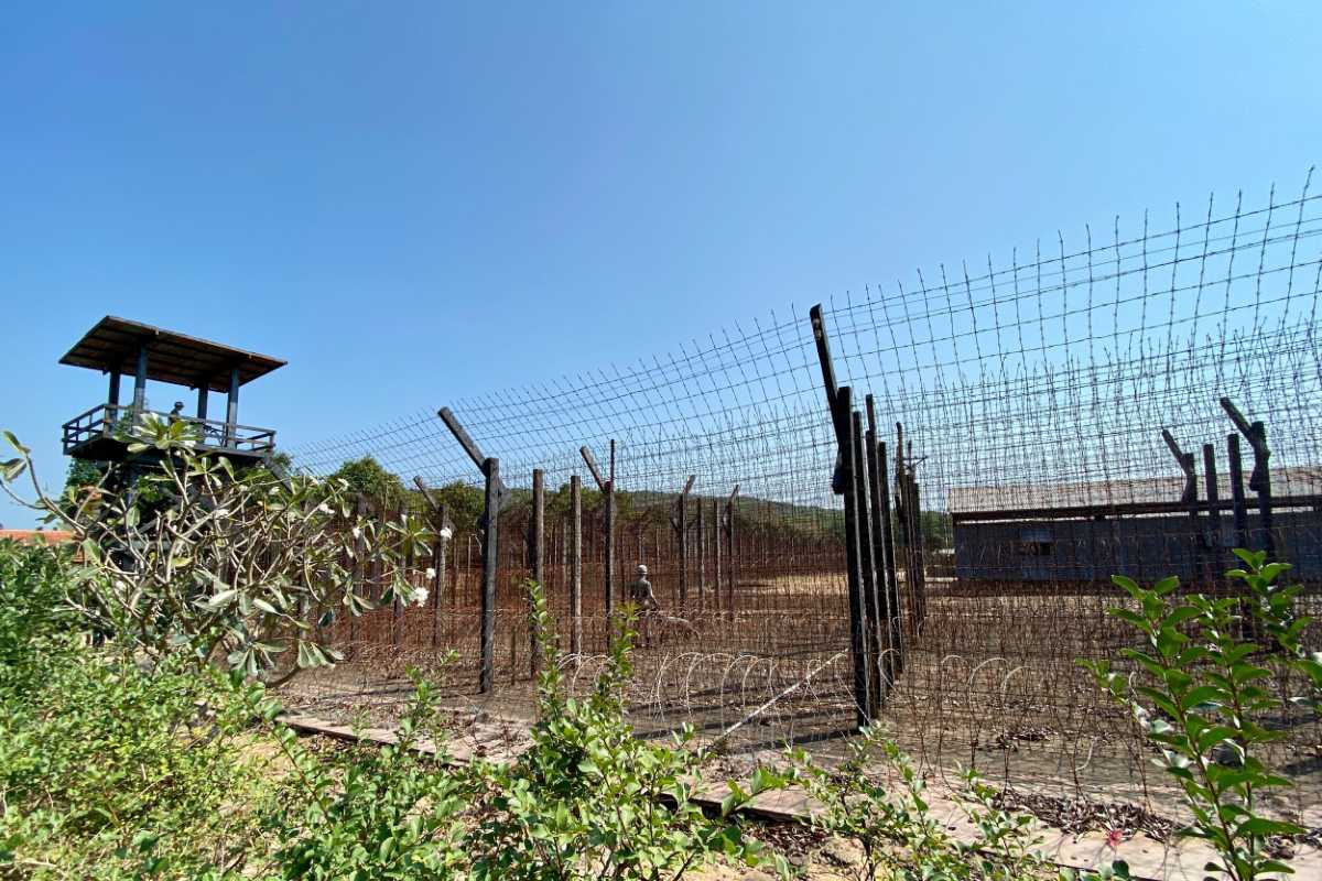 Fenced-in area of Coconut Tree Prison Phu Quoc, bordered by tropical vegetation and distant buildings.